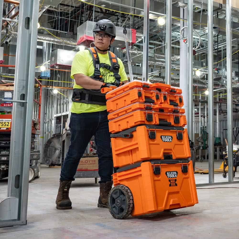 a construction worker in safety gear moves a stack of orange klein tools storage boxes on wheels inside a building under construction.